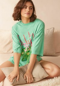Mätovo zelené oversized tričko z mäkkej bavlny, so farebným kvetinovým grafickým dizajnom. Model sedí na svetlo béžovej pohovke s vankúšom.