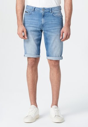 Shorts en denim bleu clair avec un ourlet retroussé, dotés de poches avant et d'une fermeture éclair, portés avec des baskets blanches sur un fond uni.