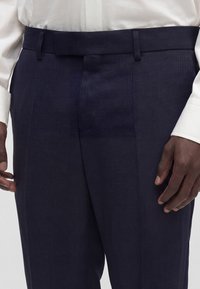Gros plan d'une personne portant un pantalon de costume texturé bleu marine foncé avec des passants de ceinture et une chemise blanche à manches longues rentrée.