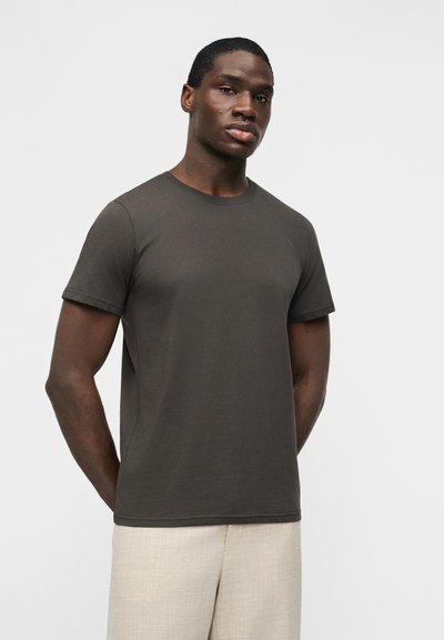 Νέος άνδρας με σκούρο δέρμα, φορώντας μια απλή σκούρα γκρι κοντομάνικη μπλούζα και ανοιχτό μπεζ παντελόνι, στέκεται μπροστά σε λευκό φόντο.