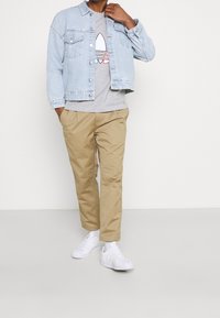 Ljust blå denimjacka över en grå t-shirt med grafisk design, kombinerad med khakibyxor och vita sneakers.
