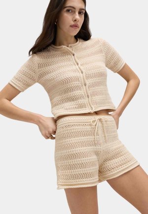 Kvinde iført matchende beige strikket kortærmet knappet top og shorts med snøre og indviklede hulmønstre.