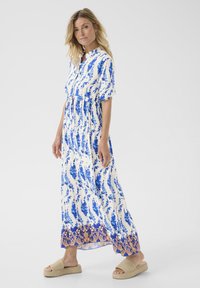 Maxi sukienka w białym kolorze z niebieskim kwiatowym wzorem, krótki rękaw, zapinana na guziki z ozdobnym dołem w pomarańczowe akcenty. Noszona z beżowymi klapkami.