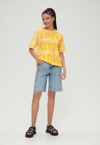 Żółta koszulka tie-dye z dużym napisem, połączona z jasnoniebieskimi szortami dżinsowymi i czarnymi sandałami. Strój charakteryzuje się luźnym krojem i casualowym stylem.