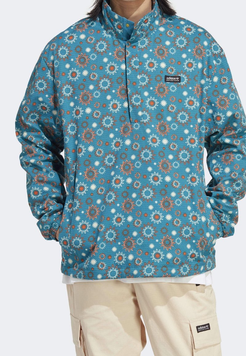 Blå tröja med solblossmönster i orange och vitt. Har en hög krage, knappslå och elastiska ärmslut.