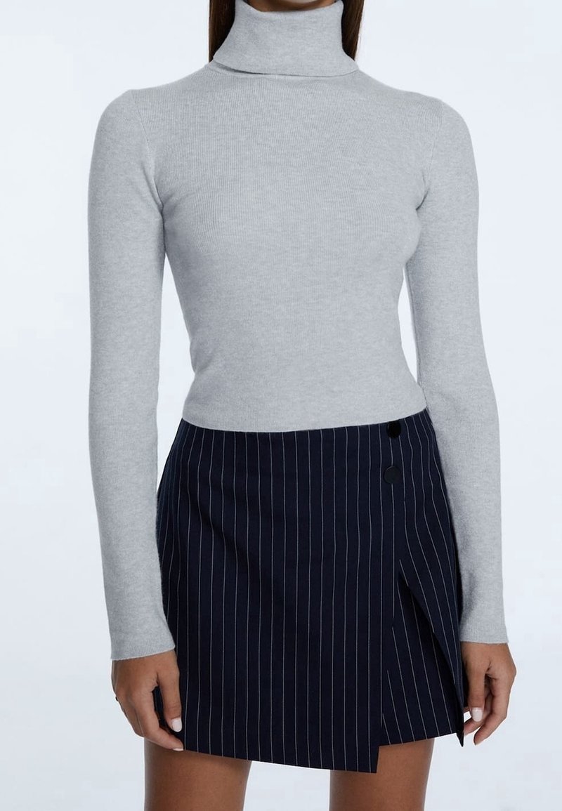 Femme portant un pull à col roulé à manches longues gris clair et une jupe bleu marine à fines rayures avec des boutons sur le côté et une fente à l'avant.