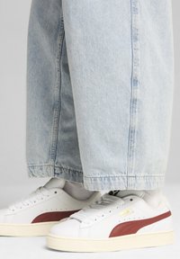 Sneakers bianchi con accenti rossi, suola in gomma testurizzata e upper in pelle liscia. Indossati con pantaloni in denim chiaro a vestibilità morbida.