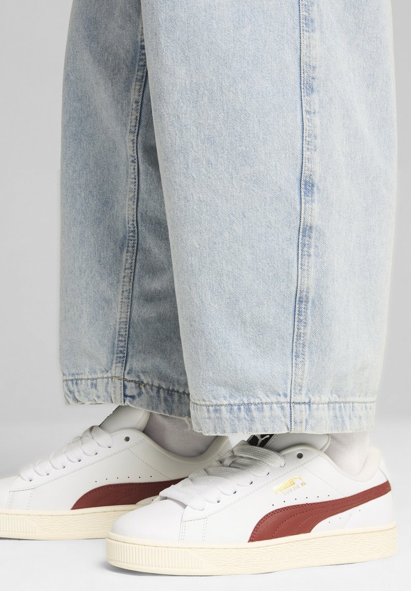 Sneakers bianchi con accenti rossi, suola in gomma testurizzata e upper in pelle liscia. Indossati con pantaloni in denim chiaro a vestibilità morbida.