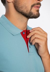 Jasnoniebieska koszulka polo z czerwonym kołnierzykiem na guziki. Kołnierzyk wyróżnia się kontrastowym kolorem i dwoma zatrzaskami do zapięcia.