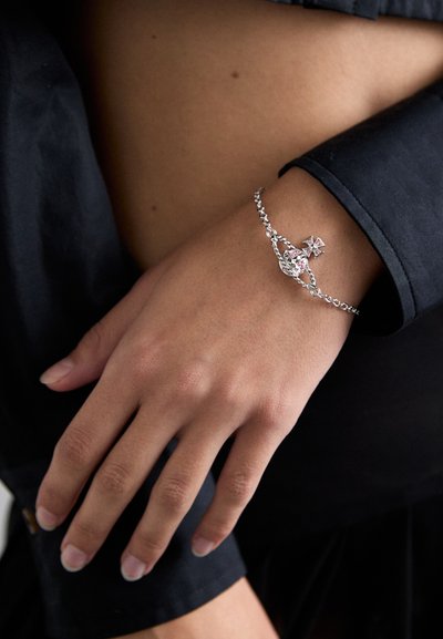 Pulseira de corrente de prata com um destaque de cristal rosa e design de cruz, usada numa wrist com unhas manicuras de forma cuidada, sobre um tecido escuro.
