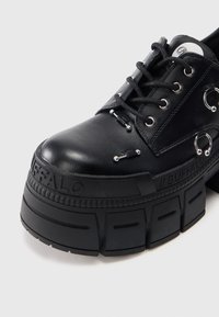 Sapato plataforma preto com sola grossa e sulcada, fecho de atacador, argolas metálicas e tachas em estilo barra, com a marca "Buffalo" na borda da sola.