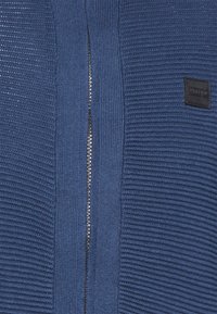 Niebieski sweter dziany z pionowym zamkiem błyskawicznym, prążkowaną teksturą po bokach oraz małą czarną metką z tłoczonymi detalami.