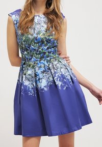 Žena oblečená v fialových šatech ke kolenům bez rukávů, zdobených modrými a bílými květinovými vzory, pózuje s jednou rukou ohnutou za zády.