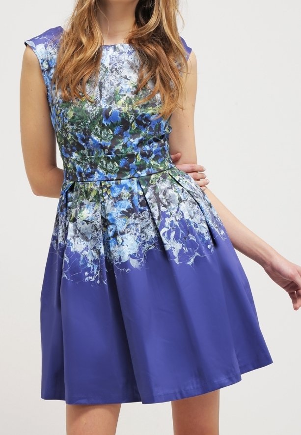 Žena oblečená v fialových šatech ke kolenům bez rukávů, zdobených modrými a bílými květinovými vzory, pózuje s jednou rukou ohnutou za zády.