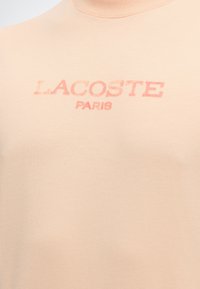 Φωτεινό μπεζ T-shirt με ξεθωριασμένο κόκκινο κείμενο "LACOSTE PARIS" τυπωμένο στο στήθος.