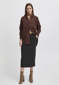 Chemise marron à boutons ample sur un col roulé beige, associée à une jupe crayon noire et des bottines brunes à bout pointu.