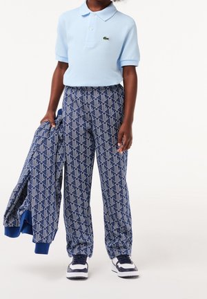 Polo bleu clair, pantalon bleu marine à motif géométrique blanc, et une veste assortie tenue dans une main. Baskets blanches avec des accents foncés.