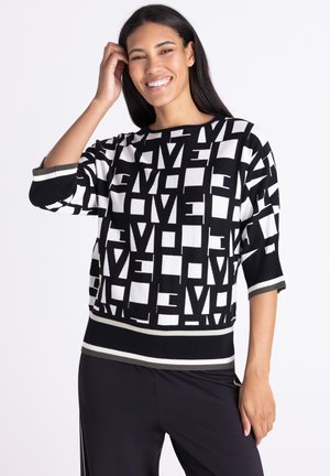 Lachende vrouw met lang donker haar draagt een zwart-witte trui met abstracte lettervormen en een zwarte broek.