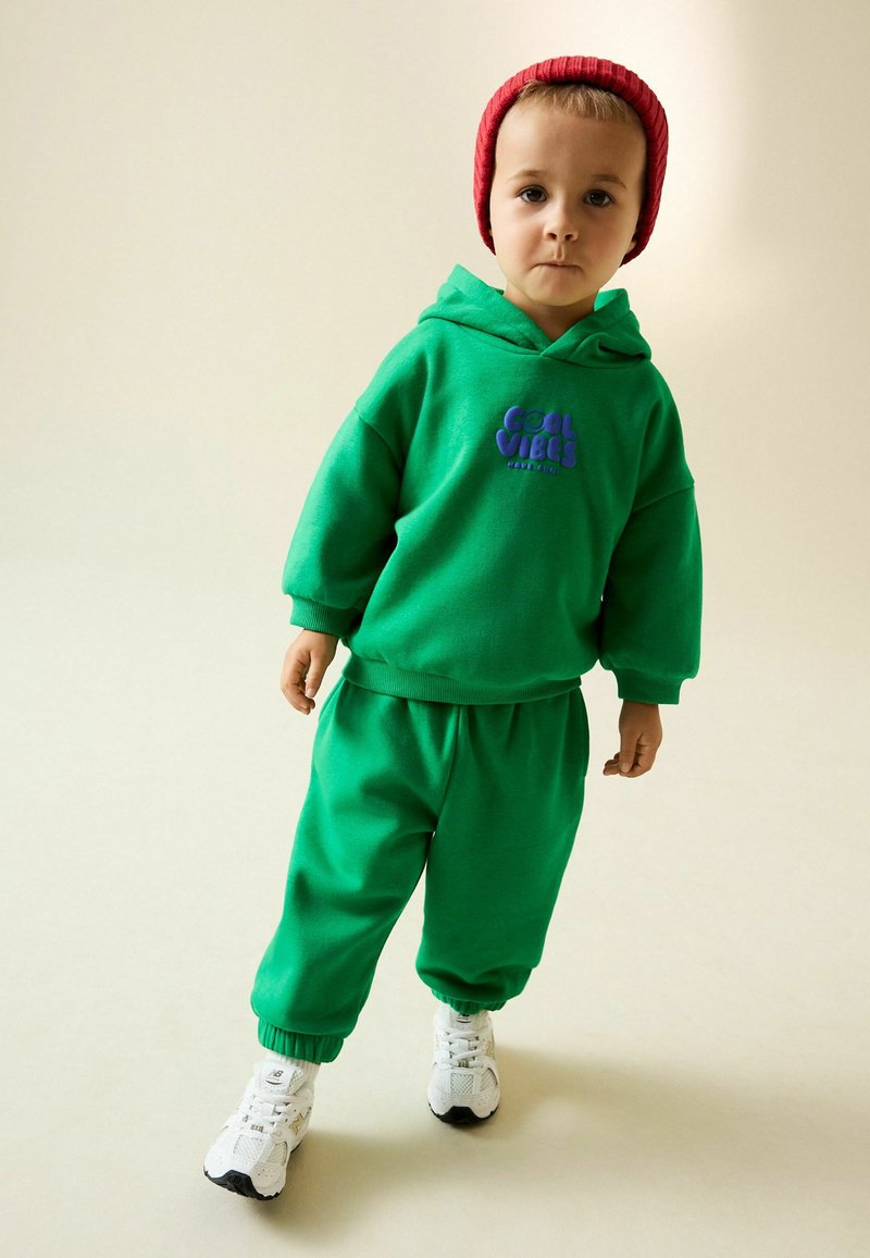 Zöld pulóver "Cool Vibes" felirattal lilában, hozzá illő melegítőnadrággal párosítva. A szettet egy piros kötött sapka és fehér sportcipők egészítik ki.