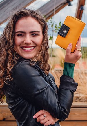 Yellow wallet with a textured finish and black closure featuring a circular logo. Model wears a black leather jacket and green sweater cuffs.