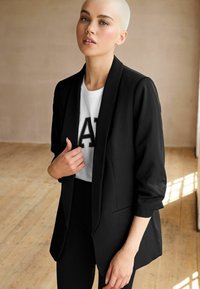 Svart blazer av smidig tyg, skräddarsydd passform med sjalkrage, ihopparad med en vit t-shirt med fet svart text.