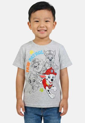 Jeune garçon souriant portant un t-shirt gris Paw Patrol avec des impressions de personnages colorées et en noir et blanc, et un jean bleu sur fond blanc.