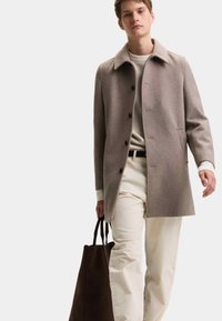 Abrigo de mezcla de lana marrón con un amplio cuello y botones negros, combinado con un suéter crema y pantalones crema. Sosteniendo un bolso tote marrón oscuro.