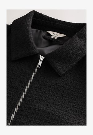 Schwarze strukturierte Jacke mit glattem Innenfutter, ausgestattet mit einem silbernen Reißverschluss und einem klassischen Kragen, beschriftet mit "NEXT" und Größe "M".