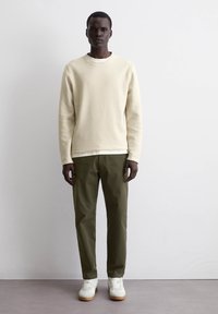 Hellbeige strukturierter Sweatshirt, olivgrüne Hose und weiße Sneaker. Einfaches, modernes Design mit Rundhalsausschnitt und subtilen Details.