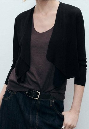 Femme portant un cardigan noir court par-dessus un haut gris foncé à encolure ronde, un jean foncé avec une ceinture noire, et des bagues en argent à la main droite.
