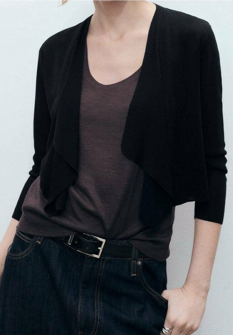Femme portant un cardigan noir court par-dessus un haut gris foncé à encolure ronde, un jean foncé avec une ceinture noire, et des bagues en argent à la main droite.