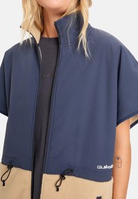 Navyblauwe ritsjack met korte mouwen, fleece onderstuk in beige, verstelbare trekkoorden en logodetail aan de zoom.