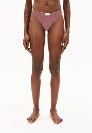 Vrouwelijk model staat blootsvoets in mauve hoge onderbroek met een wit merkje op de tailleband, tegen een witte achtergrond.
