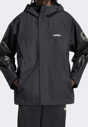 Personne portant une veste à capuche noire Adidas avec les mains dans les poches et un pantalon noir, logos Adidas visibles sur la poitrine et le pantalon.