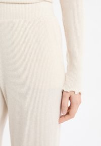 Kremowa dzianina żebrowana z fakturowanym wzorem, zaokrąglonymi mankietami z falistym brzegiem oraz dopasowaną sylwetką w talii.