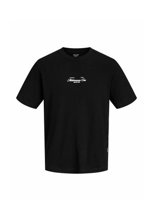 Nastolatek ubrany w czarny T-shirt z nadrukiem, beżowe szorty z kieszeniami, kremowe skarpetki z napisem i białe trampki, stojący na jednolitym tle.