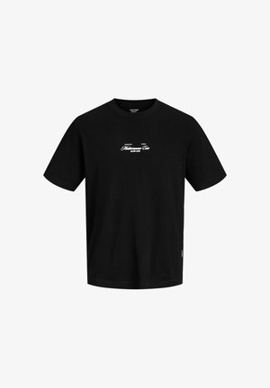 Teenage dreng iført en sort t-shirt med print, beige shorts med lommer, cremefarvede sokker med tekst og hvide sneakers, der står mod en ensfarvet baggrund.