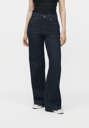 Vrouw in een donkerblauwe high-waisted jeans met wijde pijpen en zwart-witte sneakers, staand tegen een effen lichte achtergrond.