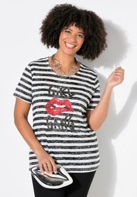 Camiseta a rayas en blanco y negro con un gráfico de labios en lentejuelas rojas y texto negro, combinada con un clutch metálico plateado y pantalones negros.