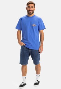 Blaues Baumwoll-T-Shirt mit einem kleinen Grafikdesign auf der Brust, kombiniert mit Jeansshorts, weißen Socken und schwarzen Skate-Schuhen.