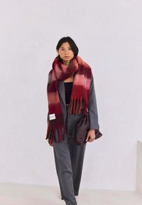 Écharpe surdimensionnée rouge et marron dégradé avec des extrémités à franges, portée sur une tenue sombre, complétée par un sac marron.