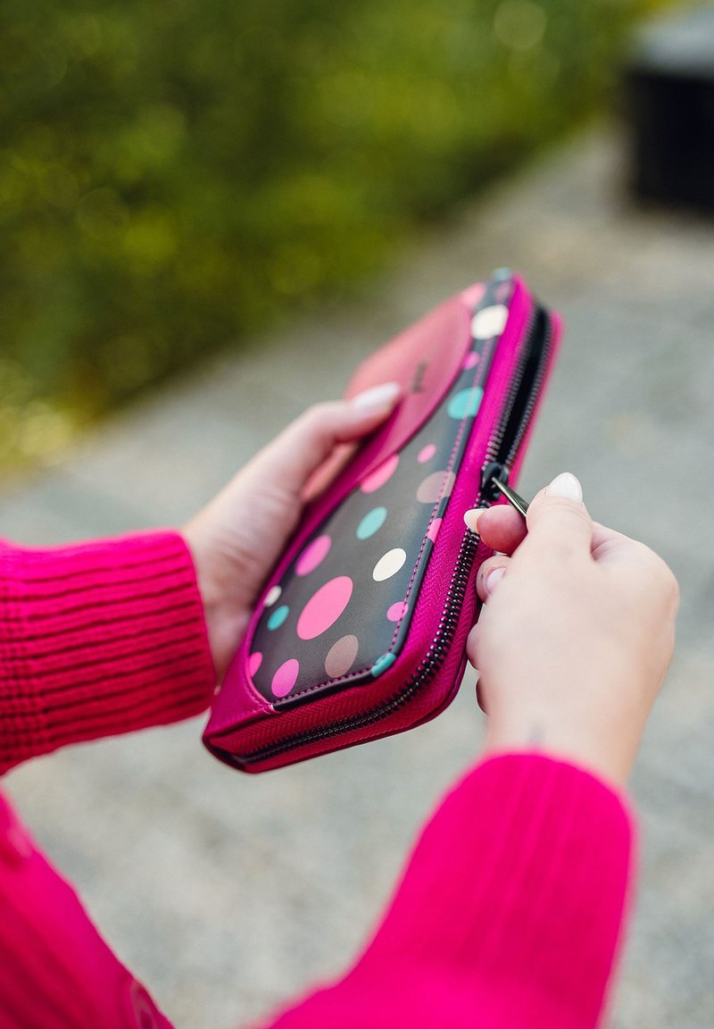 Zippered wallet with a black surface featuring multicolored polka dots, framed in bright pink material. Hand holds the zipper pull.