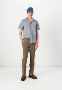 Camicia azzurra a maniche corte con bottoni e un motivo, abbinata a pantaloni khaki e scarpe in pelle marrone. Un cappellino blu completa il look.