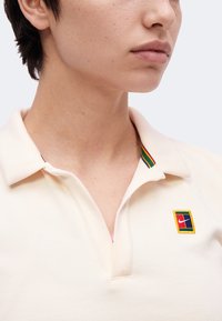 Gros plan d'une personne portant un polo crème avec un col rayé multicolore et un petit écusson brodé sur la poitrine.