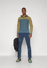 Άνδρας που στέκεται φορώντας μπλε και ελαιοπράσινο ρουχισμό και παντελόνι για προστασία από το κρύο, κρατώντας μαύρο σακίδιο, με αντίστοιχα μπλε και πράσινα μποτάκια πεζοπορίας.