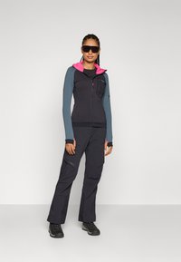 Schwarze und graue Reißverschlussjacke mit einer rosa Kapuze, geschichtetes Design. Kombiniert mit schwarzen Cargo-Hosen und schwarzen Wanderschuhen.