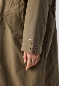 Ελαιοπράσινο ύφασμα παλτό με κουμπωτό μανίκι, ορατή τσέπη και μια μικρή λογότυπο λεπτομέρεια. Η υφή φαίνεται ομαλή και ελαφρώς τσαλακωμένη.