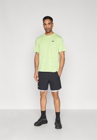 Πράσινο αθλητικό μπλουζάκι με κοντά μανίκια και λογότυπο, συνδυασμένο με μαύρο σορτς με λευκά περιμετρικά σχέδια. Μαύρα αθλητικά παπούτσια και κάλτσες ολοκληρώνουν την εμφάνιση.