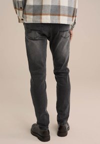 Jeans de color gris oscuro con un corte ajustado, que presentan bolsillos traseros y una textura desgastada. Combinados con zapatos marrones de cordones, vistos desde detrás.