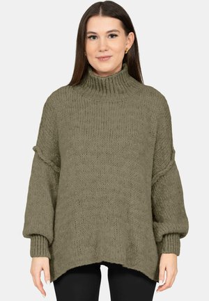 Frau mit langen dunklen Haaren, die einen übergroßen olivgrünen Rollkragenpullover und schwarze Hosen trägt, steht vor einem einfarbigen Hintergrund.